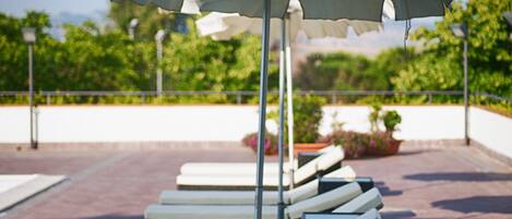 A row of sun loungers with an umbrella, a railing, and greenery in the background.