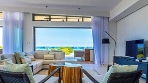 A modern living room with a sofa, chairs, a TV, and a view of the ocean. - Manta Cove by Horizon Holidays (Black River)