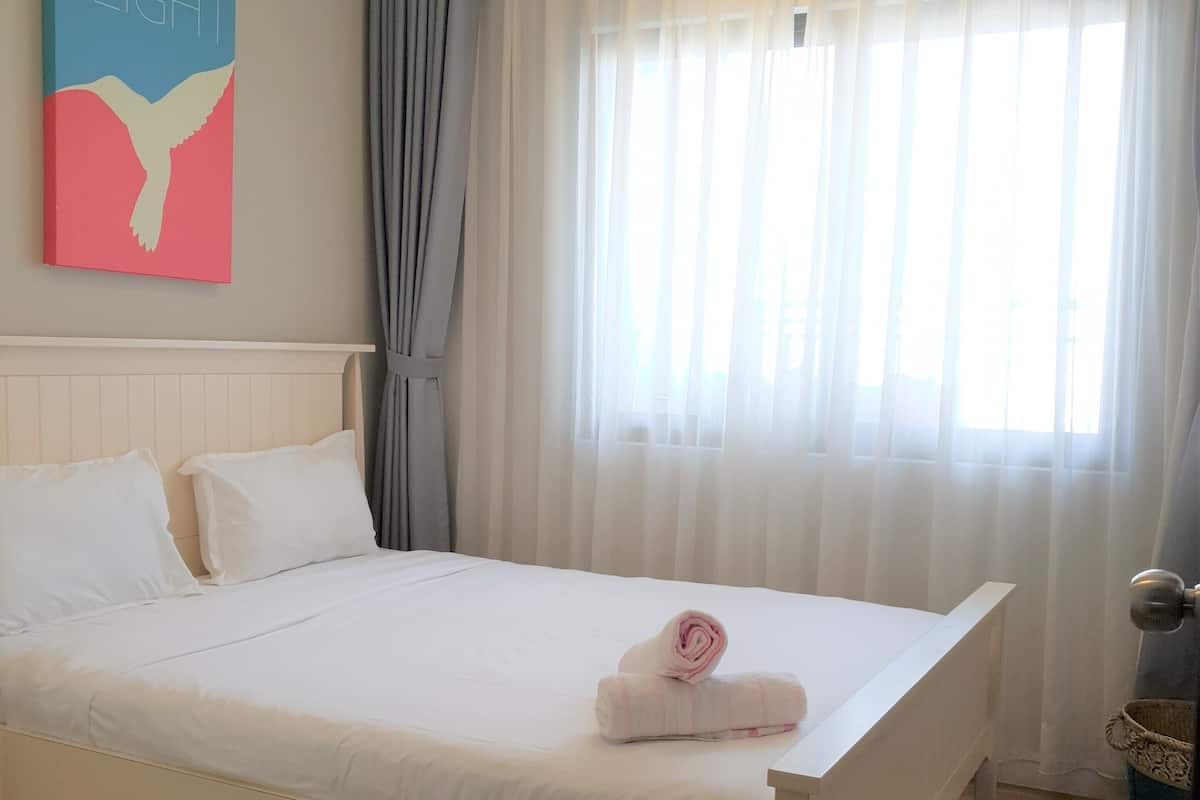 A neatly made bed with white linens, a wall-mounted artwork depicting a bird, and a window with sheer curtains.