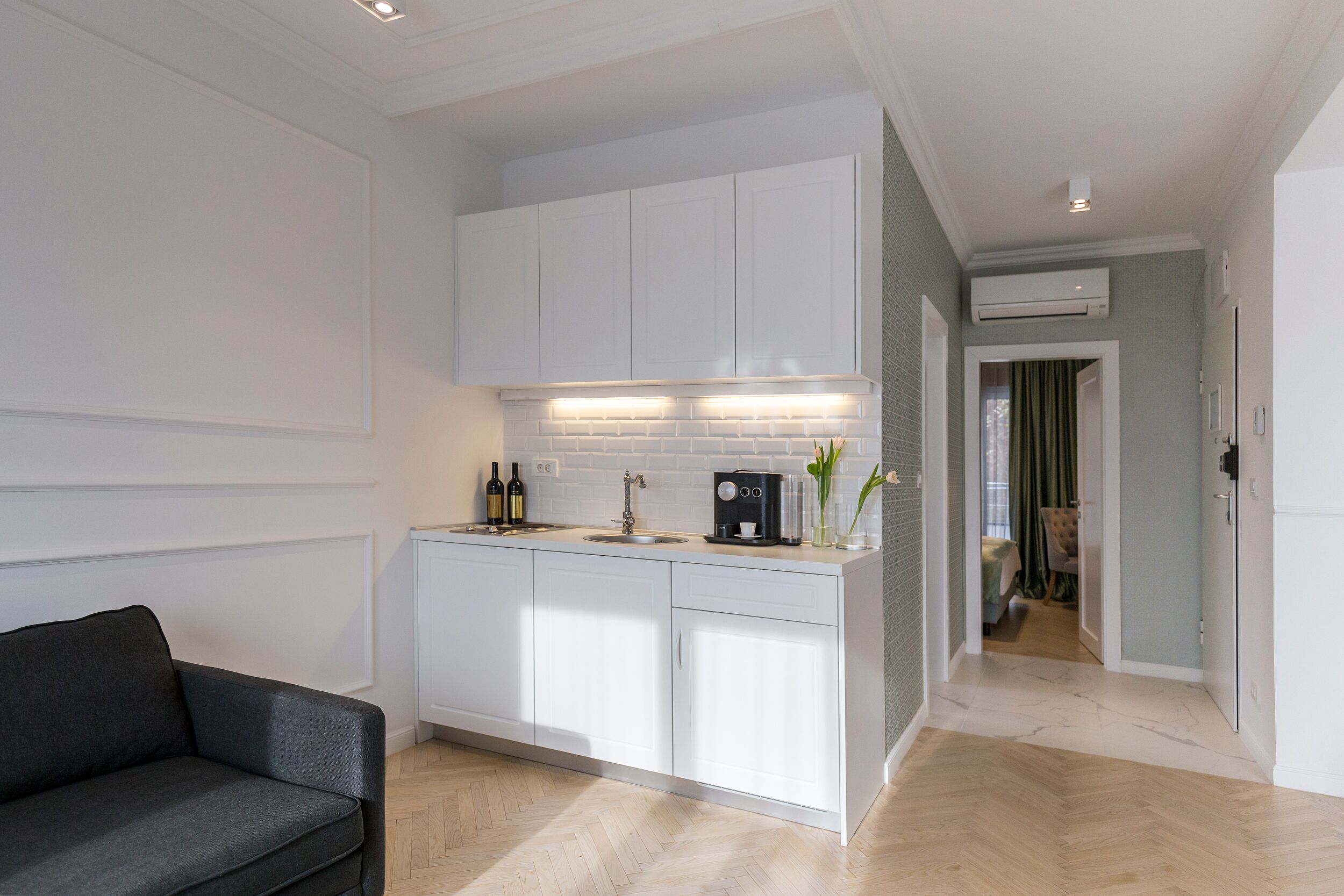 A modern kitchen with white cabinetry, a built-in oven, and a coffee machine. There is a sofa in the foreground and a dining area visible through an open door.