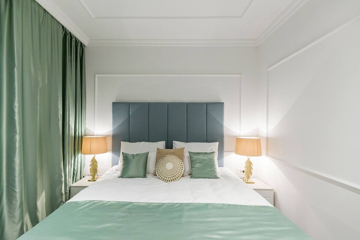 A neatly arranged hotel room with a large bed, green curtains, and two bedside lamps.