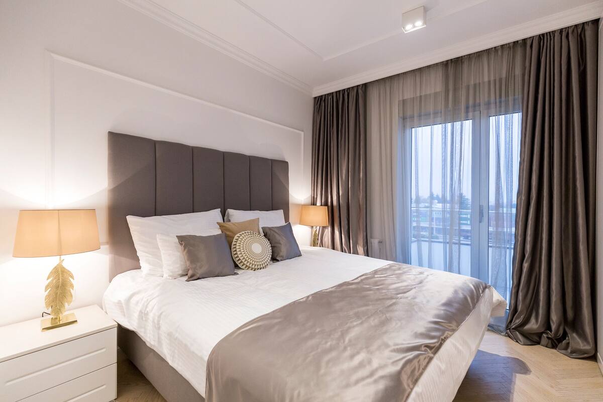 A modern hotel room with a large bed, two bedside tables with lamps, a view of the city through large windows, and a cityscape visible outside.