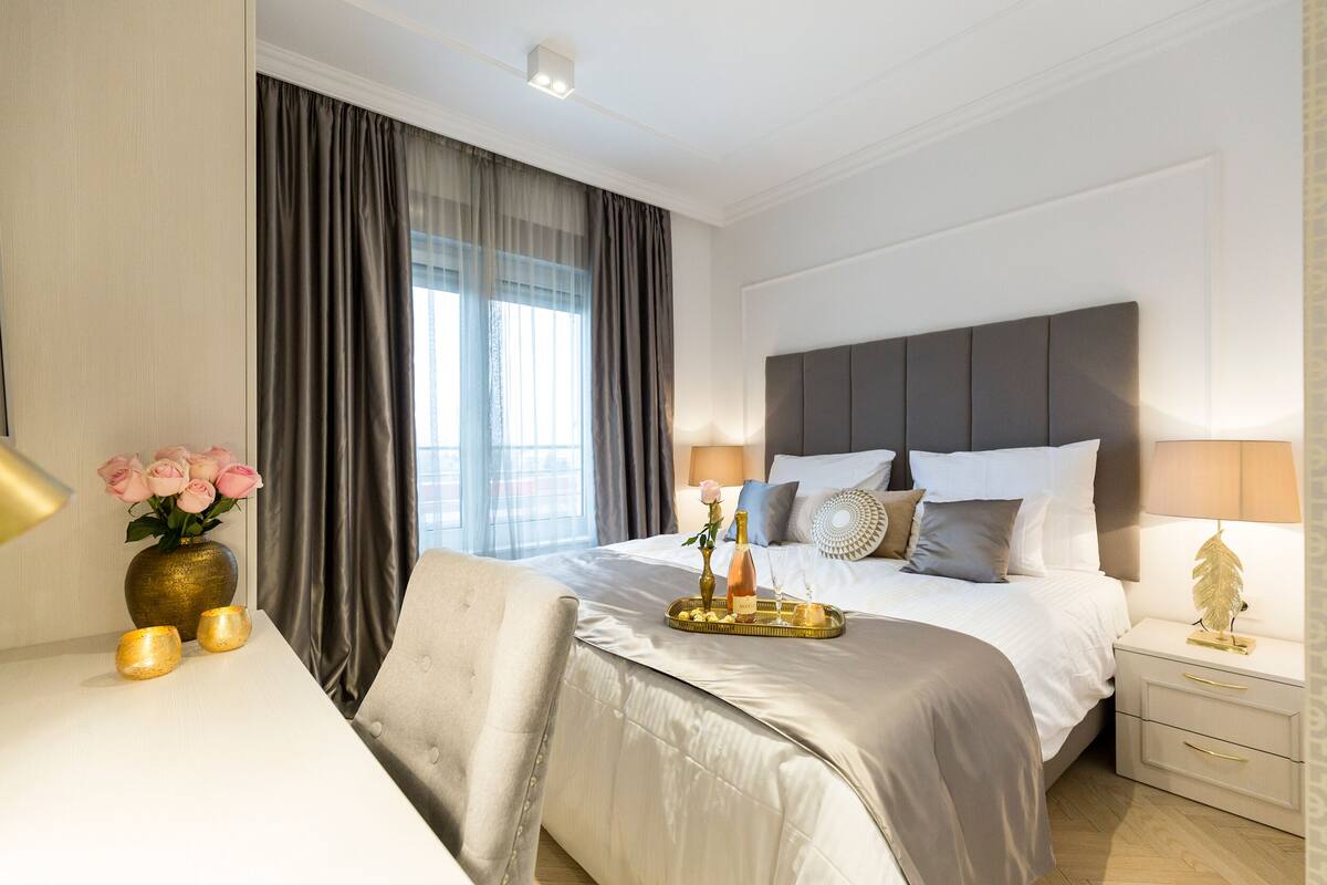 A modern hotel room with a large bed, a bedside table with a lamp, a vase with flowers, and a tray with candles.