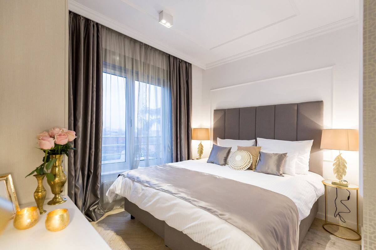 A hotel room with a large bed, a bedside table with a lamp, a vase with flowers, and a view of the city through the window.