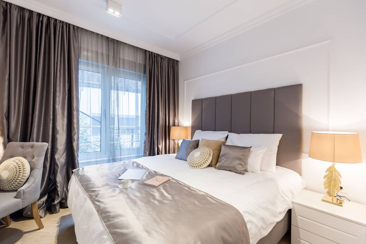 A modern hotel room with a large bed, a bedside table with a lamp, a chair, and a view of the city through large windows.
