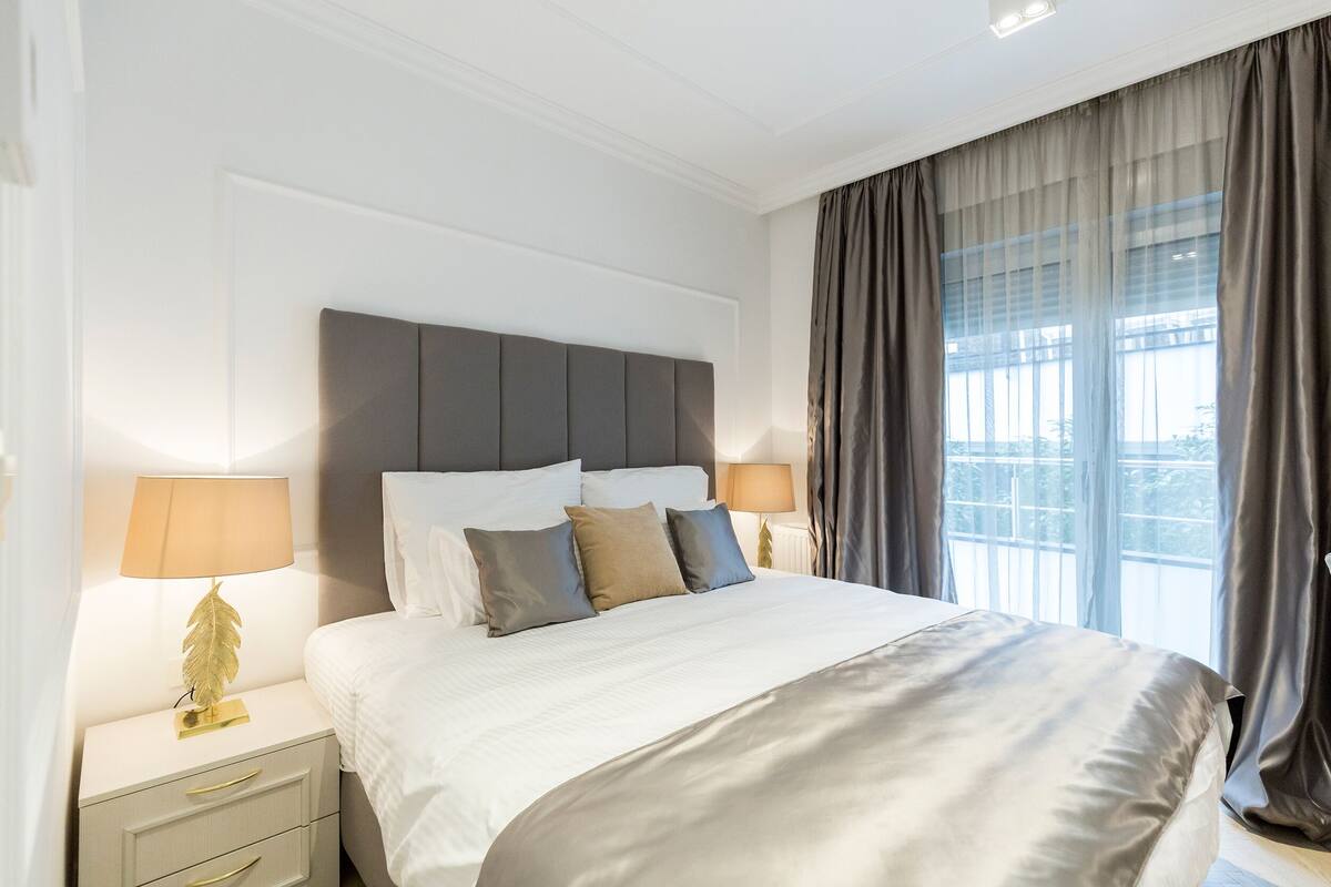 A neatly made bed with a grey headboard, two bedside tables with lamps, and a large window with curtains.