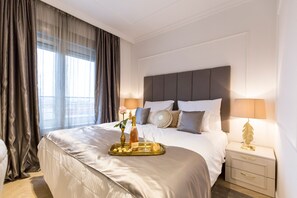 A neatly made bed with a tray of snacks, a bottle, and glasses, a bedside lamp, and a window with curtains. - MM House - Adults Only (Zagreb)