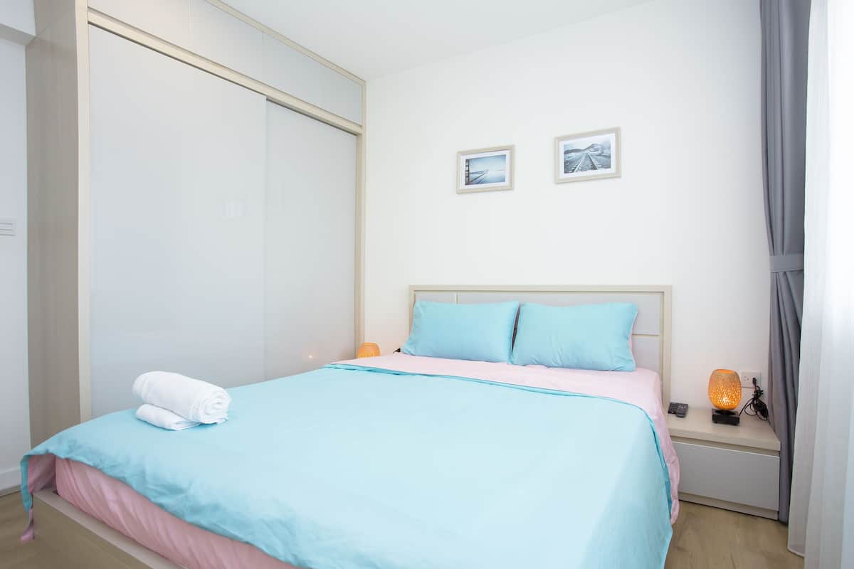 A neatly made bed with a blue and pink bedspread, a white towel, and a bedside lamp.
