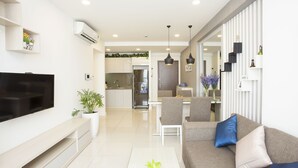 A modern living room with a flat-screen TV, a sofa with cushions, a coffee table with a plant, and a kitchen in the background. - Ami Splendid Rooftop Garden (Ho Chi Minh City)