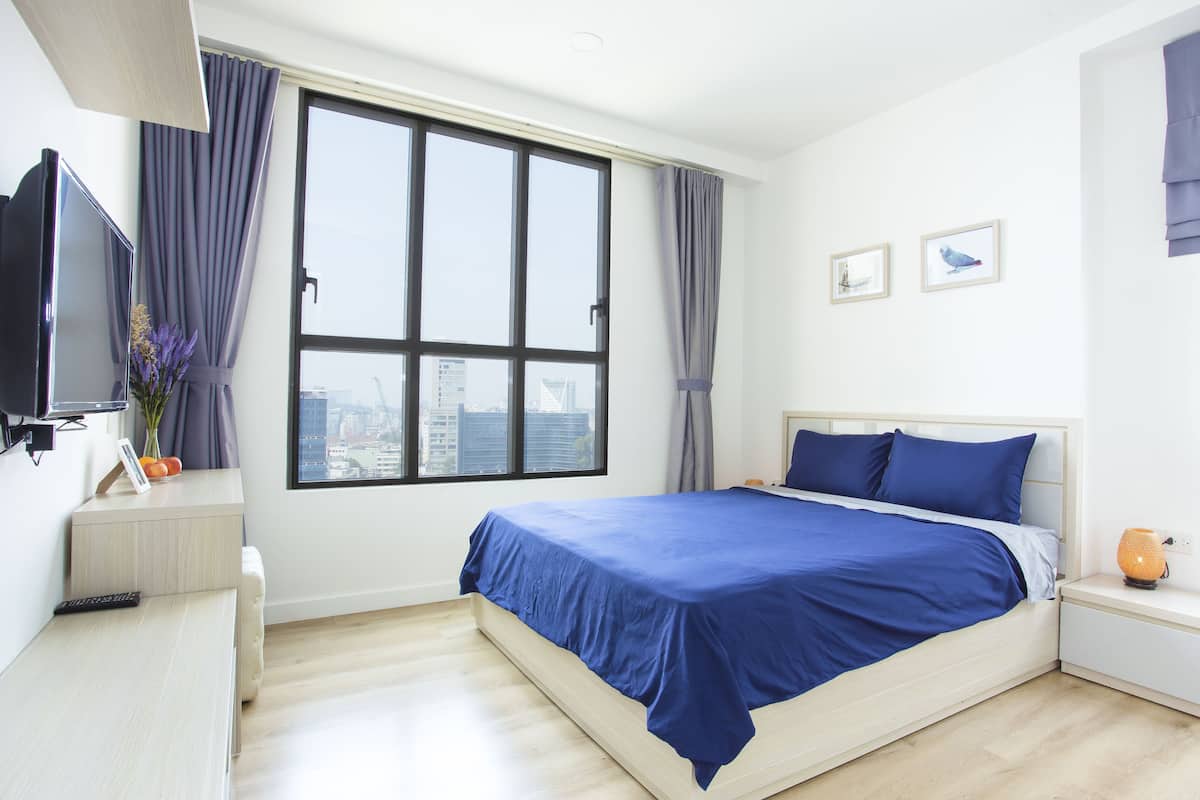 A modern bedroom with a large window, a bed with blue bedding, a bedside table with a lamp, and a TV on the left.