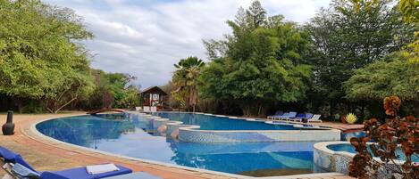 Una piscina con mosaicos azules, rodeada de sillones de descanso y palmeras.