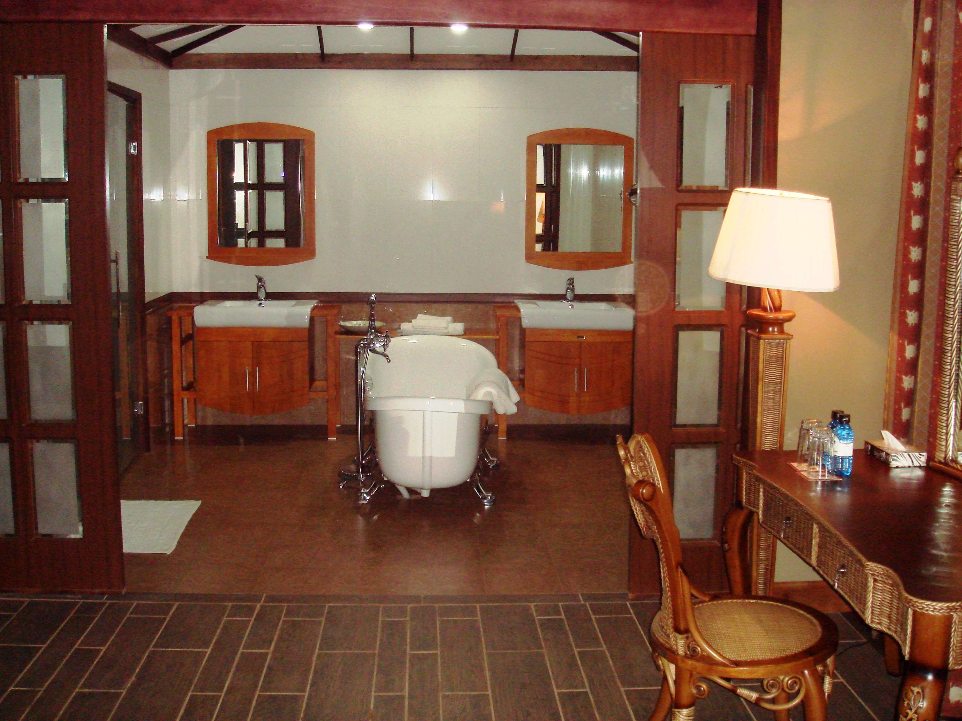 A bathroom with a bathtub, two sinks, a mirror, a chair, and a table with a lamp and toiletries.