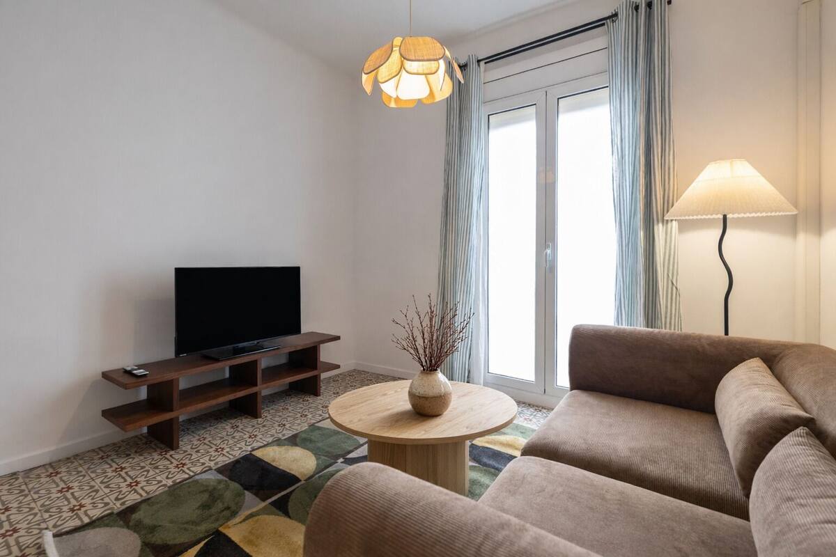 Un salón moderno con un televisor de pantalla plana sobre un mueble de madera, una mesa de centro redonda con un florero, un sofá marrón y una lámpara.