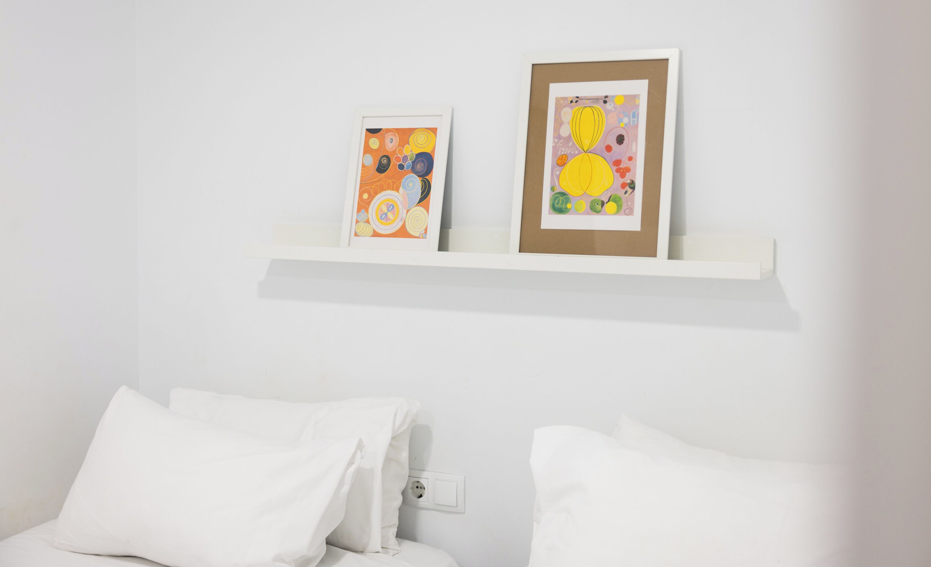Un dormitorio con una cama blanca, almohadas blancas y una pared blanca. Hay un estante blanco con dos obras de arte enmarcadas.