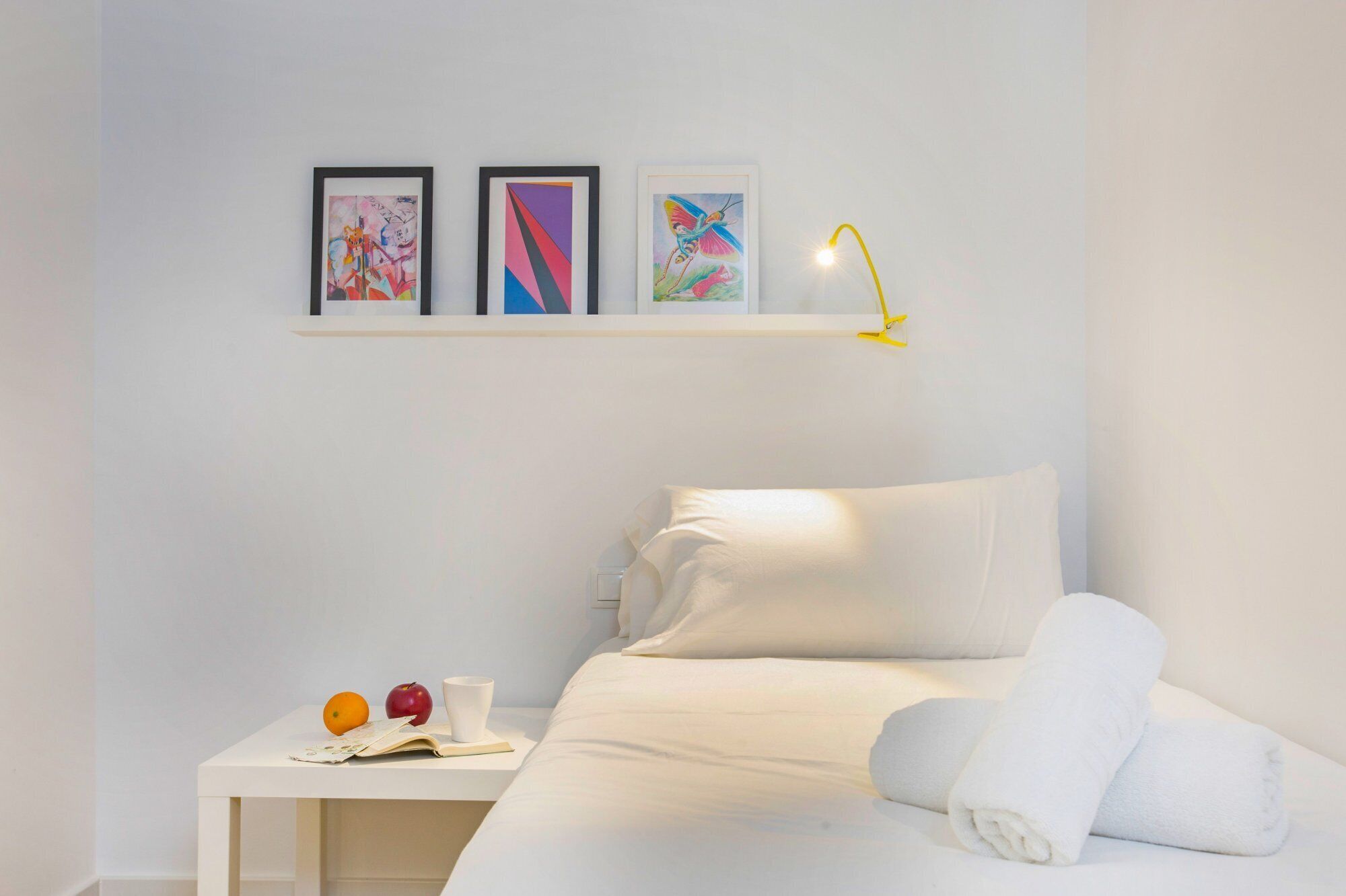 A hotel room with a bed, a nightstand with a book, an apple, and a cup. There are three framed artworks on the wall above the bed.