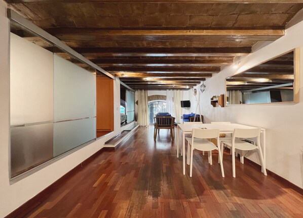 Un pasillo amplio con suelo de madera, un comedor con sillas blancas y un espejo grande en la pared derecha.