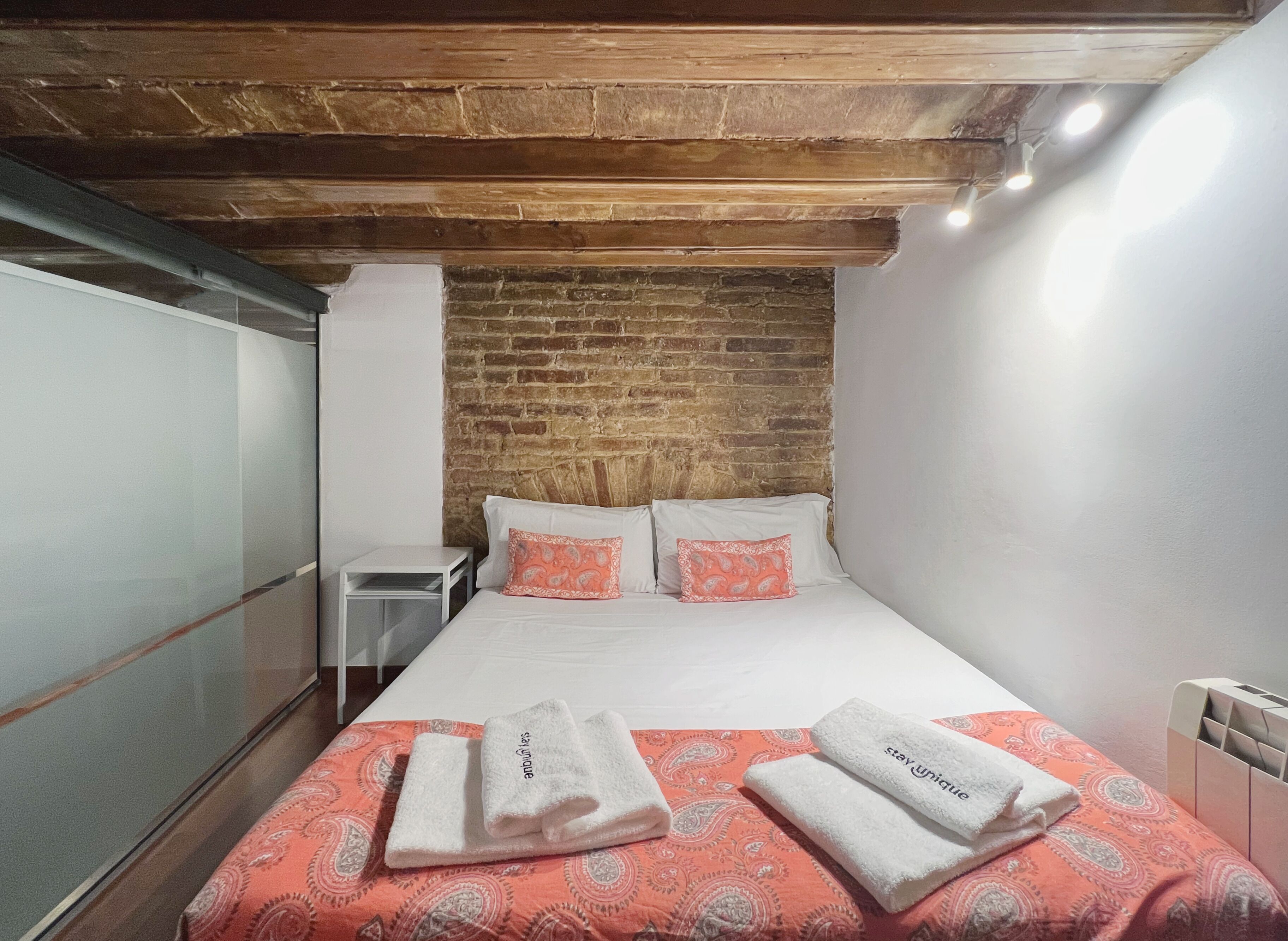Une chambre d’hôtel avec un mur en briques, un lit avec du linge de lit blanc, deux oreillers, une couvre-lit rouge à motifs et deux serviettes avec du texte imprimé dessus.