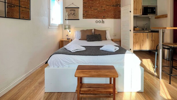 Une chambre moderne avec un grand lit, un banc en bois, une table de chevet avec une lampe, un mur en briques et un coin cuisine visible à travers une porte ouverte.