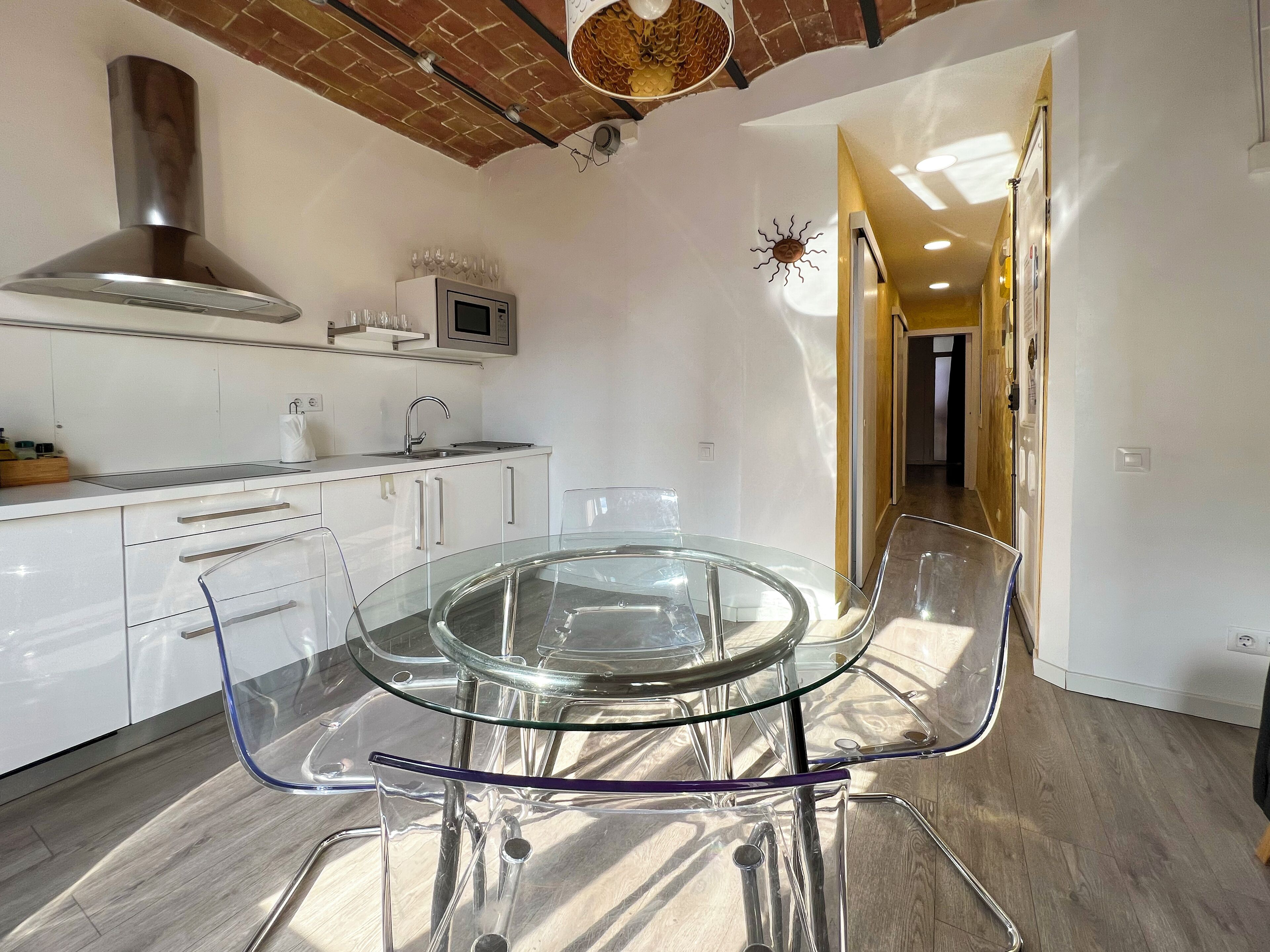A modern kitchen with a glass dining table, white cabinetry, and a brick ceiling.