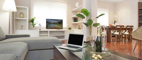 A modern living room with a sofa, a glass coffee table, a laptop, and a dining area with chairs and a table.