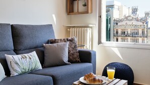 A modern living room with a grey sofa, a wooden coffee table with croissants and orange juice, and a view of a building with unique architecture. - Stay U-nique Apartments Rambla Catalunya (Barcelona)