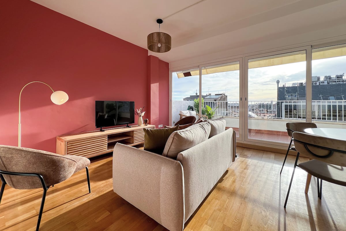 Una sala de estar moderna con un televisor grande, un sofá, una mesa de comedor y un balcón con vista a la ciudad.