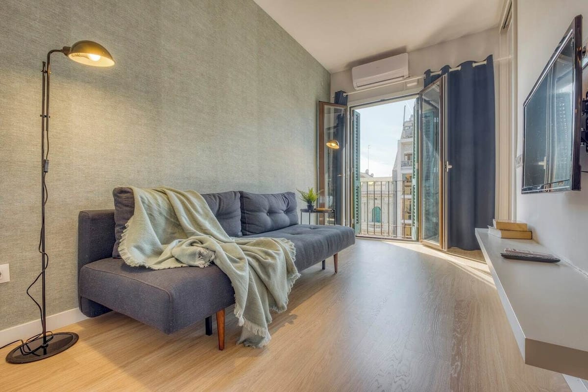 Una sala de estar moderna con un sofá gris, una lámpara de pie y vistas a la ciudad a través de grandes puertas de vidrio.