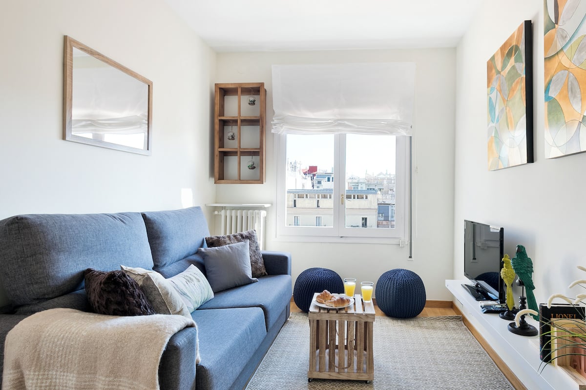 Una sala de estar moderna con un sofá azul, una pequeña mesita de madera con cosas para el desayuno, un ventanal con vistas a edificios y un televisor montado.