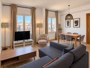 A modern living room with a flat-screen TV, grey sofas, a wooden coffee table, and a dining area with a wooden table and chairs.