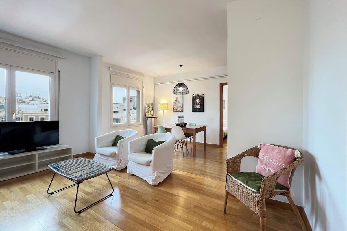 Una sala de estar moderna con un televisor de pantalla plana, dos sillones blancos, una silla de mimbre, una mesa de comedor con sillas y vistas a la ciudad a través de amplios ventanales.