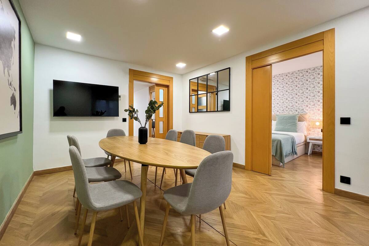 Un comedor con una mesa redonda de madera, seis sillas grises, un jarrón negro con plantas, un televisor de pantalla plana y un espejo con marco de madera.