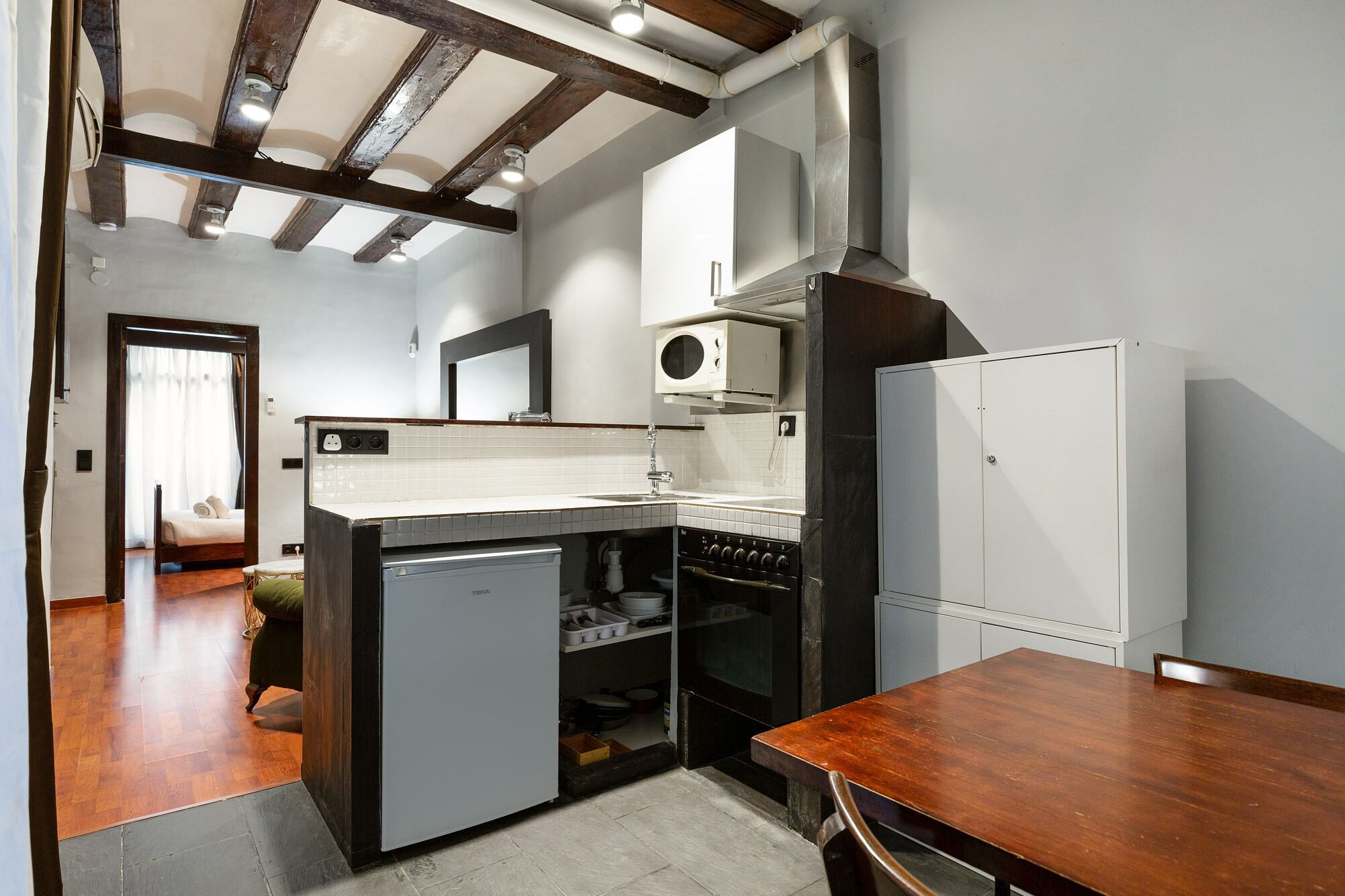 A compact kitchen with a built-in dishwasher, a microwave, and a stove. There is a wooden dining table and a bed visible in the adjacent room.
