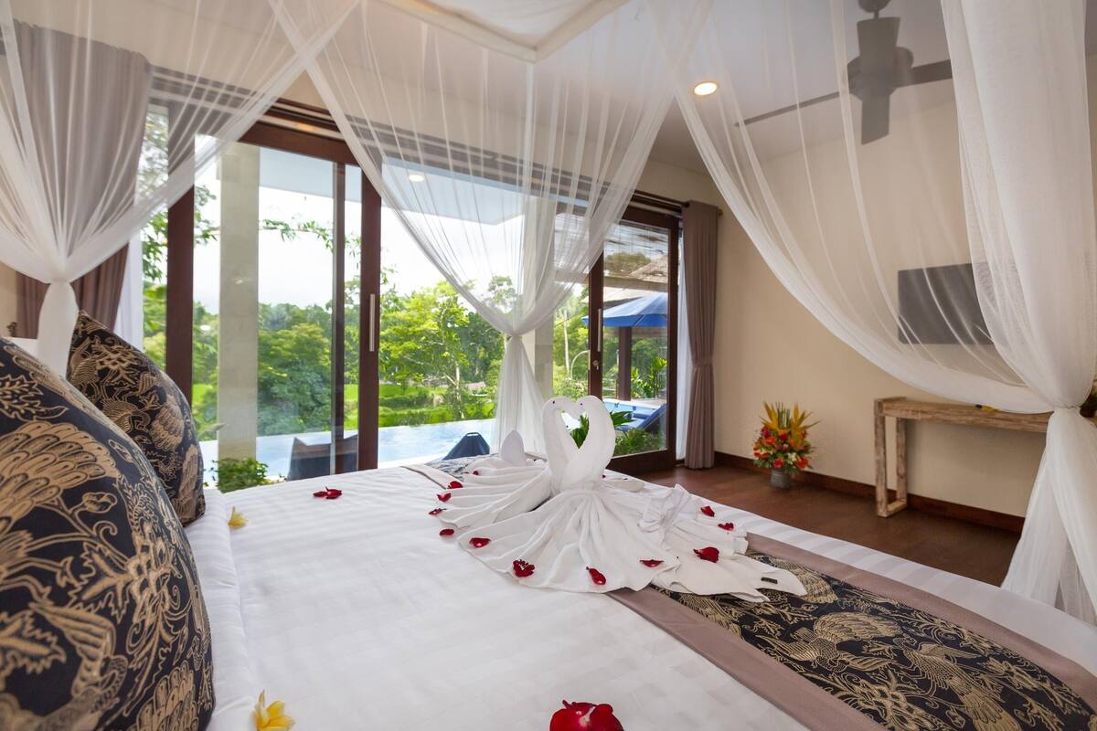 A hotel room with a bed adorned with a swan-shaped arrangement of white and red rose petals, a canopy bed, and a view of lush greenery outside.