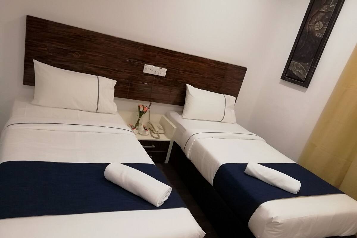 A hotel room with two beds, each with a blue bedspread and white linens, a wooden headboard, a bedside table with a phone and a vase of flowers, and a wall-mounted artwork.