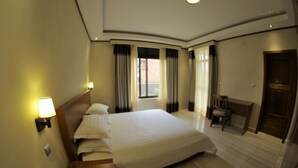 A hotel room with a large bed, a desk, a chair, a window with curtains, and a door. - Nandi Residence (Kigali)