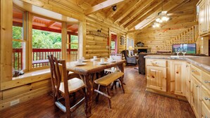 A wooden cabin interior with a dining area, kitchen, and a large window overlooking greenery.