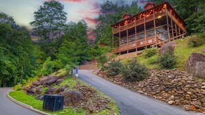 A wooden cabin with a balcony, surrounded by a forested landscape and a winding road.