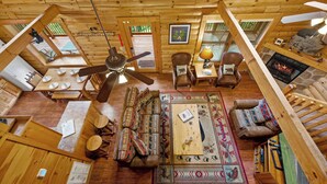 A cozy cabin interior with a wooden ceiling, a central ceiling fan, a fireplace, a sofa, a coffee table, and a dining table set for four.