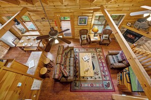 Interior acogedor de una cabaña con techo de madera, un ventilador de techo central, chimenea, sofá, mesa de centro y una mesa de comedor preparada para cuatro.
