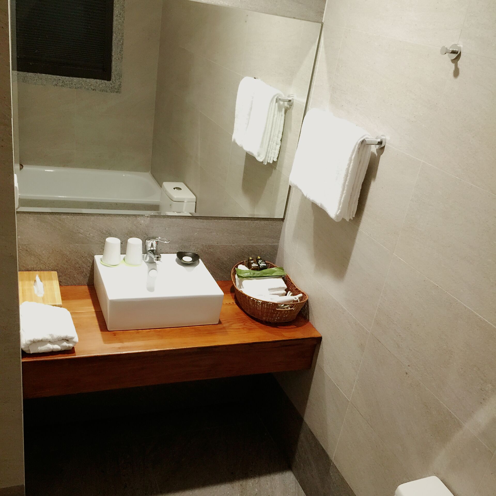 Een badkamer met een houten wastafelmeubel, een witte wastafel en een spiegel. Er is een handdoekhouder aan de muur en een mandje met toiletartikelen.