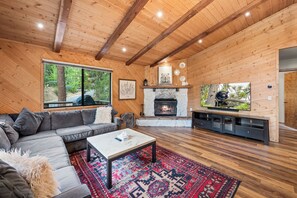 Een knus interieur van een blokhut met een stenen openhaard, een groot raam met uitzicht op groen en een gezellige zithoek.