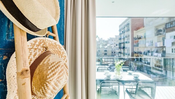 Parvekkeella on pöytä ja tuolit, ja sieltä avautuu näkymä rakennuksiin. Seinällä roikkuu hattuja.