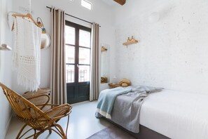 A room with a bed, a wicker chair, a door with curtains, and a white brick wall.