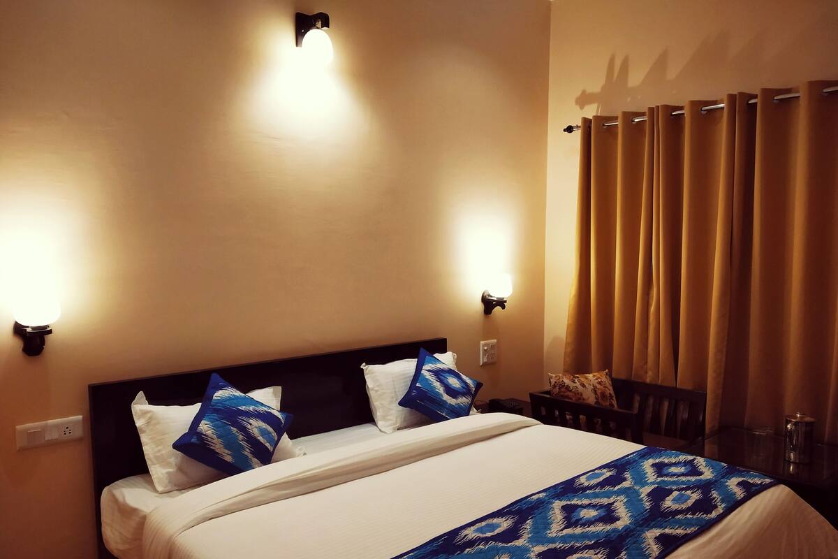 A hotel room with a bed, two wall-mounted lights, a window with curtains, and a bedside table with a lamp.