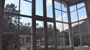 A room with large windows offering a view of a hillside with buildings and trees. - Hotel Vista (Nainital)