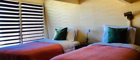 Quarto de hotel com duas camas, cada uma com colcha vermelha, uma janela com persianas e um ponto de luz fixado na parede.
