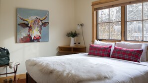 A bedroom with a large bed, a painting of a cow on the wall, a window with a view of trees, and a bedside table with a lamp and a plant. - Outpost: Alpine Cottage Three (Jackson)