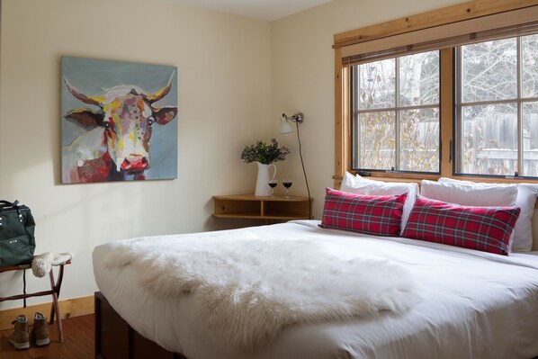 A bedroom with a large bed, a painting of a cow on the wall, a window with a view of trees, and a bedside table with a lamp and a plant. - Outpost: Alpine Cottage Three (Jackson)