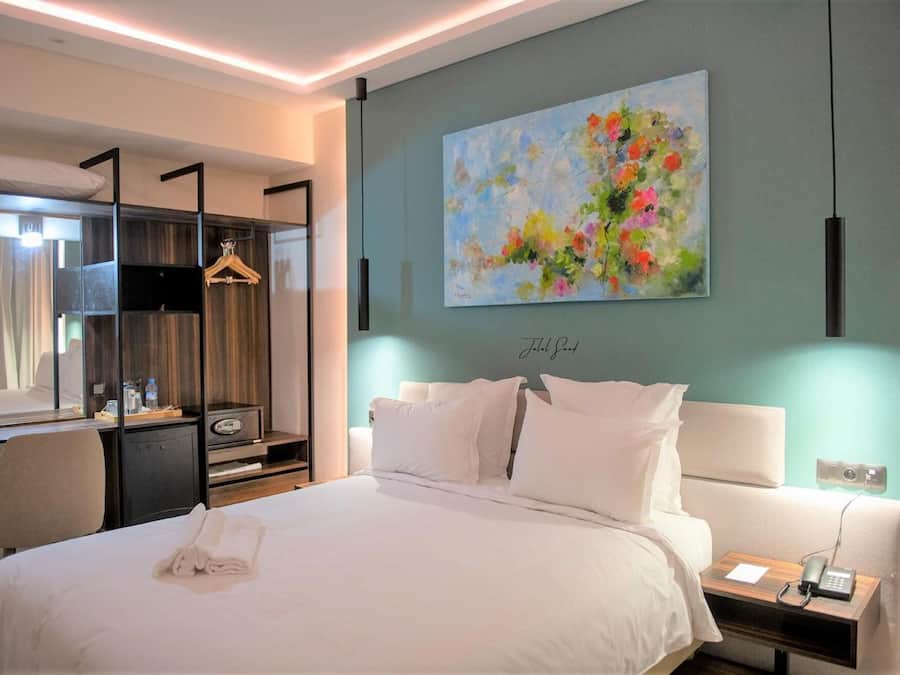 A hotel room with a bed, bedside tables, a desk with a chair, a wardrobe, and a painting on the wall.