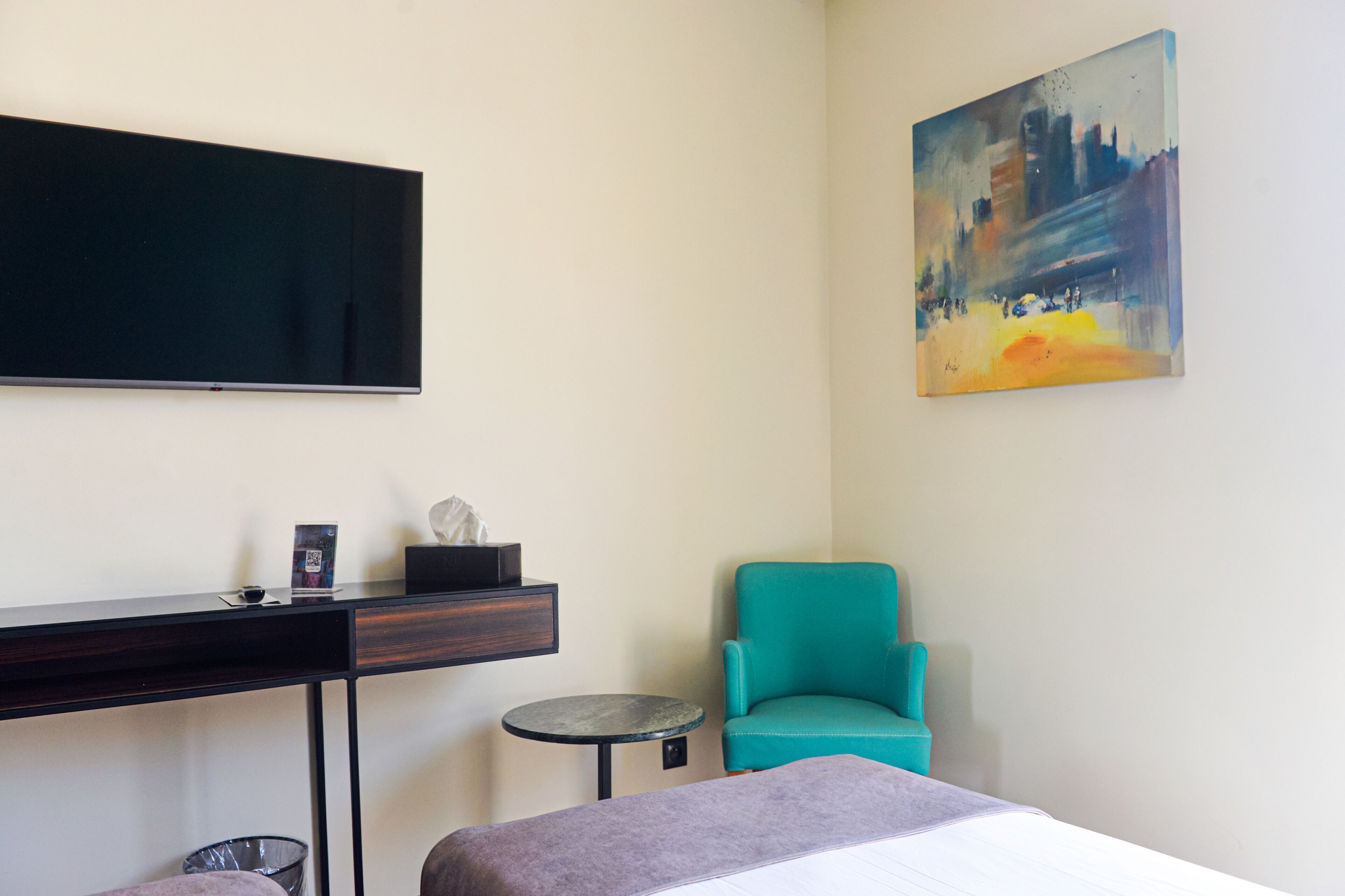 A hotel room with a bed, a desk, a chair, a small table, and a painting on the wall.
